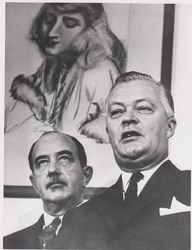Jean Lesage pose devant une oeuvre d'art représentant une femme. Un homme derrière lui lève les yeux au ciel.
