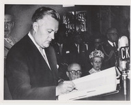 Jean Lesage signe un document. Une femme et quelques hommes assistent à la cérémonie retransmise sur les ondes de la radio